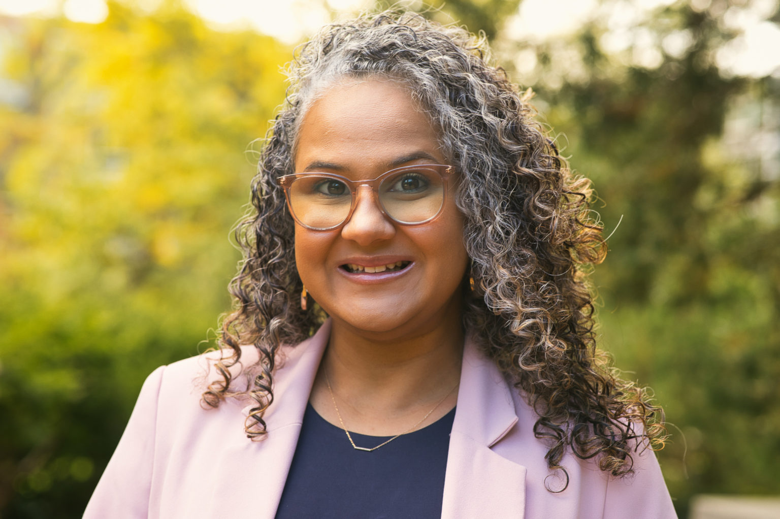 Person with gray-brown hair wearing light pink translucent glasses and a light pink blazer