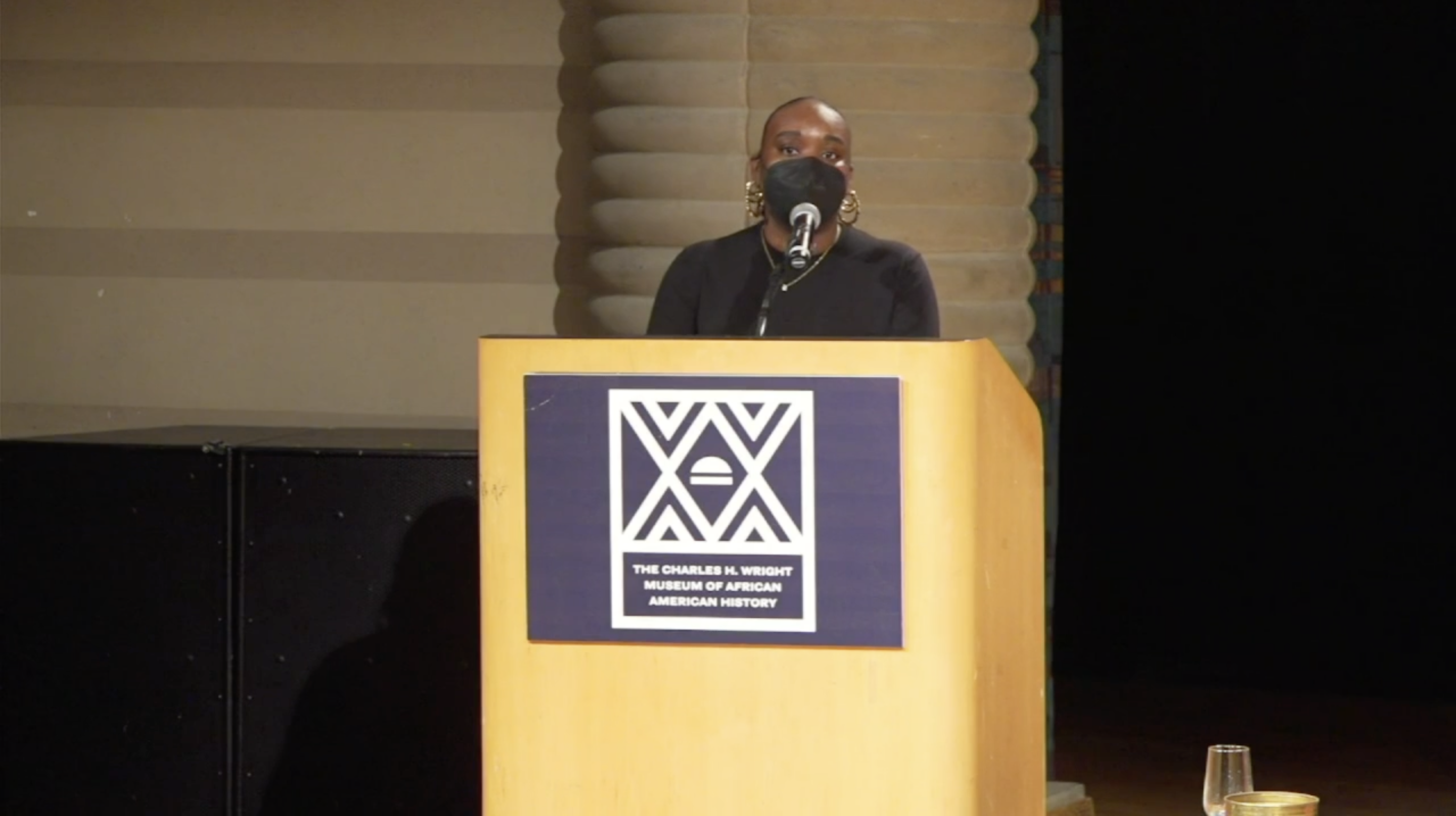 Woman speaking at a podium. She is wearing a black face mask and black top.