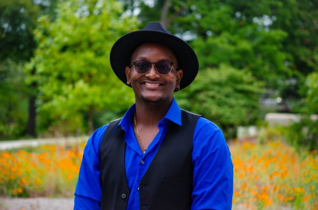 A young man in a hat, sunglasses, and blue shirt smiles in front of a field of flowers.