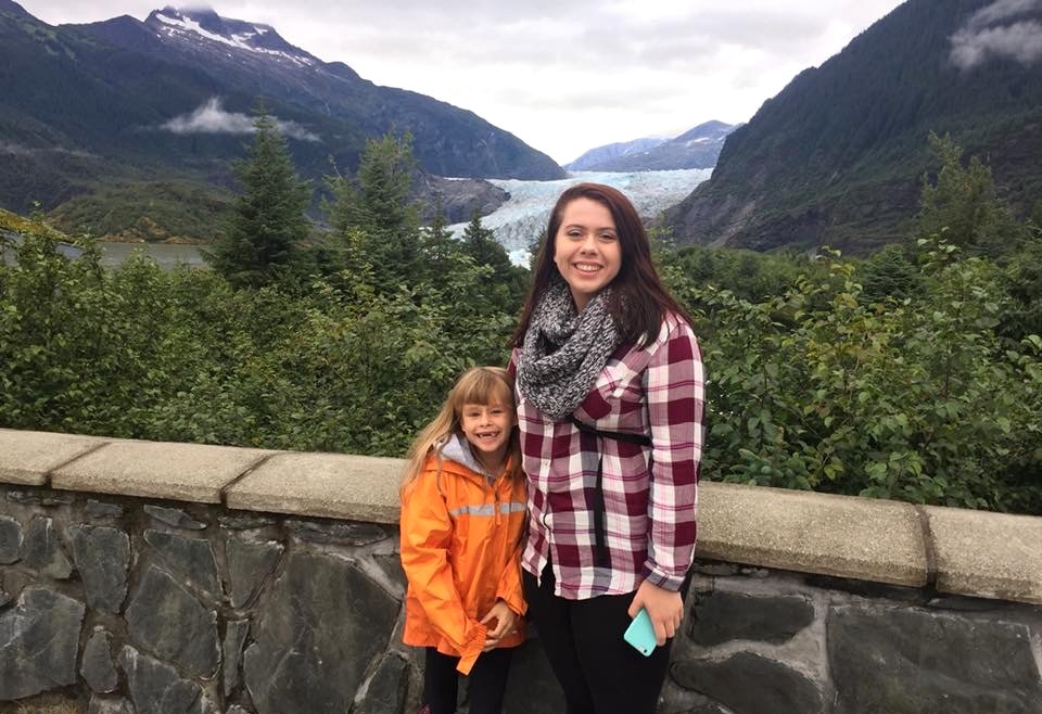 Woman wearing a red and white plaid top and scarf standing next to a child wearing an orange coat. In the background is a glacier and mountains.