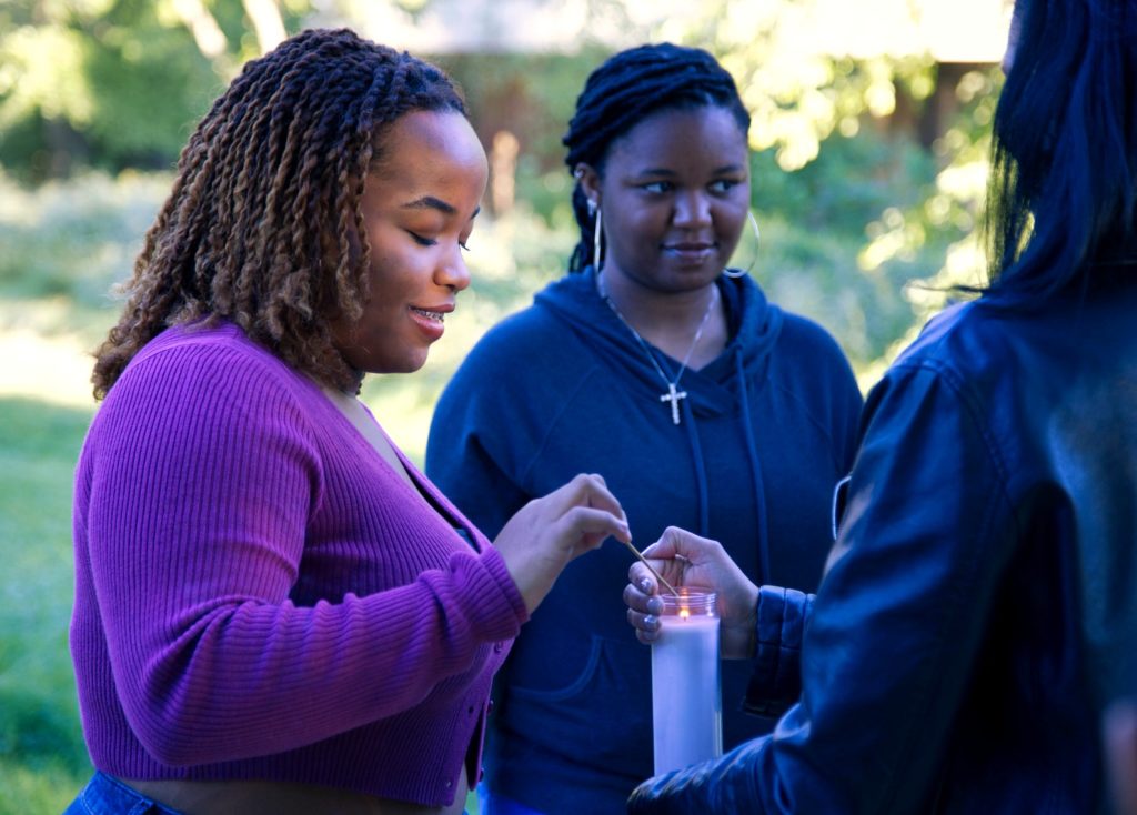 Three Black women standing next to each other. One wearing a putle long-sleeve shirt and is lighting a candle. Another is holding the candle and her face cannot be seen. Another is wearing a black sweatshirt and is wearing a necklace with a cross on it. 