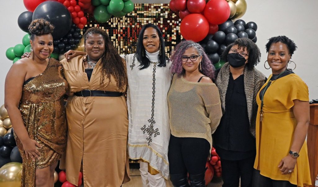 Group of six Black women standing in a row with arms around each other with red, green, black,and gold balloons in the background.