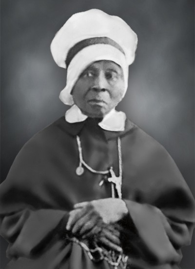 Antique photo of a woman sitting for a portrait. She is wearing a white head covering and long black clothing.