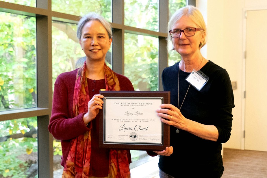 Two women stand smiling, holding an award certificate. The woman on the left wears a red sweater and scarf, while the woman on the right wears a black top and glasses. They stand in front of a large window with greenery visible outside. 