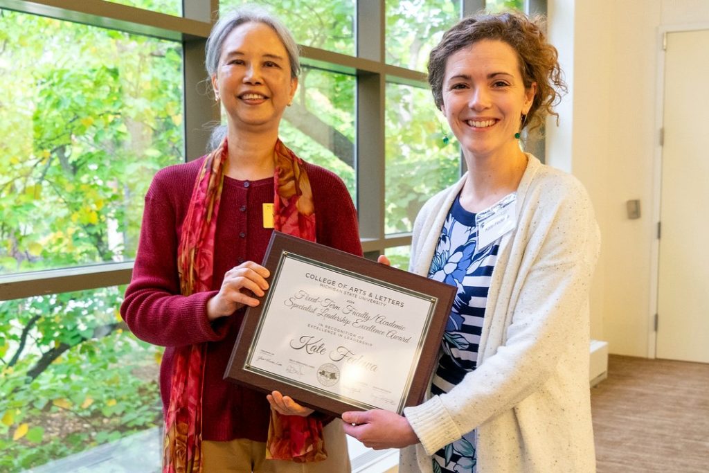 Two women stand smiling, holding an award certificate. The woman on the left wears a red sweater and scarf, while the woman on the right wears a white cardigan and blue patterned top. They stand in front of a large window with greenery visible outside. 